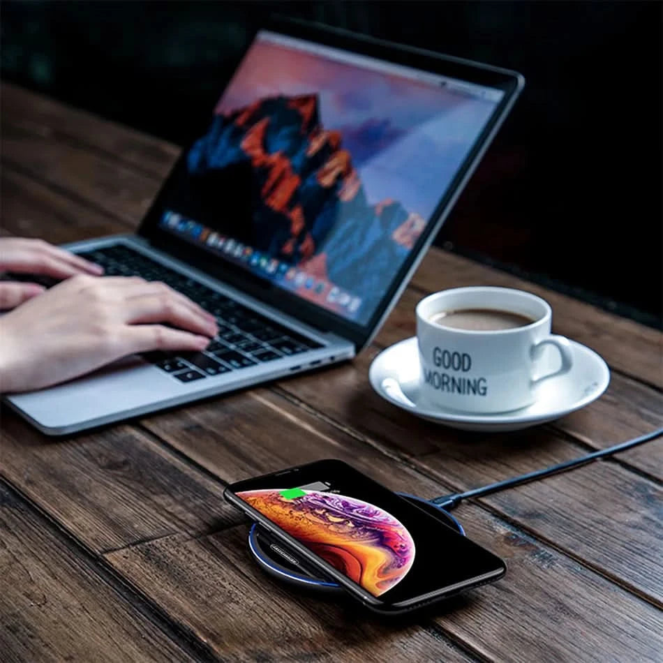 Ricarica wireless su base circolare: smartphone in carica accanto a laptop e una tazza di caffè.