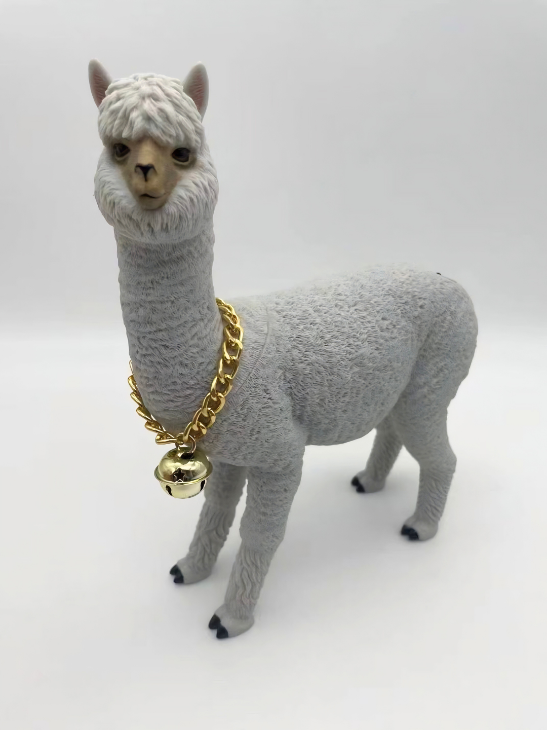 Alpaca grigia in stile cartoon con collana dorata e campanella, su sfondo bianco.